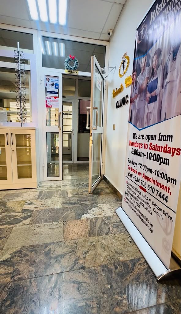 Entrance to Diplomatic Village Clinic beside a banner showing the clinic's opening hours and appointment line